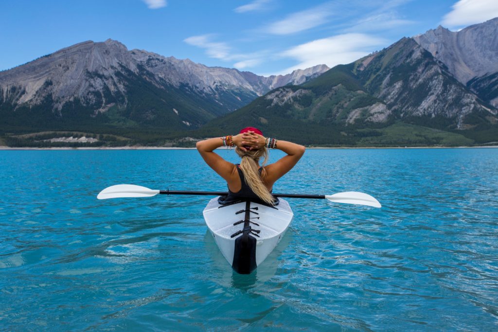 A woman wearing a tank top and red cap is practicing mindfulness in nature as she kayaks in crystal clear water by a mountain