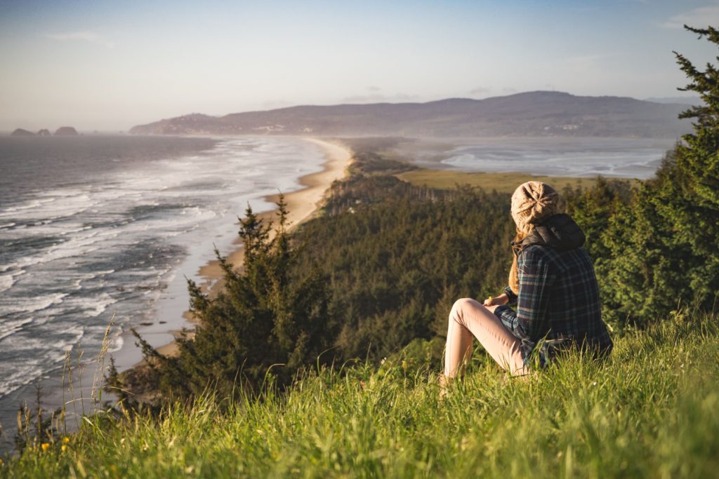 A woman in a flannel, jeans, and a knit cap is meditating in nature as she sits on a grass knoll overlooking the sea