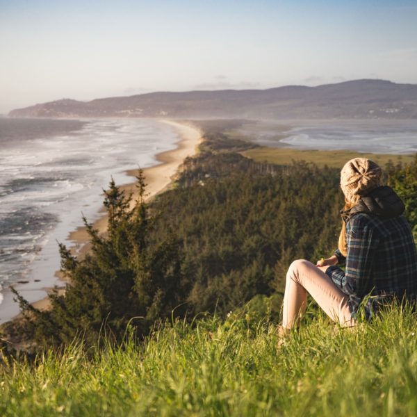 A woman in a flannel, jeans, and a knit cap is meditating in nature as she sits on a grass knoll overlooking the sea